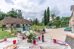 A wooden table set for dining with plates, glasses, and a bottle, surrounded by greenery and a building in the background.