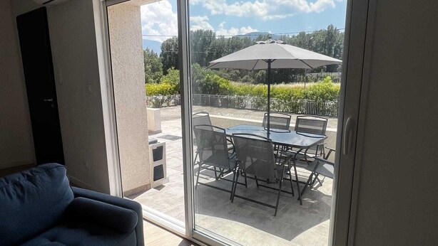 A room with a sofa, a sliding glass door leading to an outdoor patio with a table and chairs, and an umbrella. - Cosy apartment ideally located in the center of Corsica, close to hiking and beaches. (PIEDIGRIGGIO)