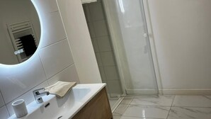 A modern bathroom with a white sink, wooden vanity, a round mirror, and a glass shower enclosure. - Cosy apartment ideally located in the center of Corsica, close to hiking and beaches. (PIEDIGRIGGIO)