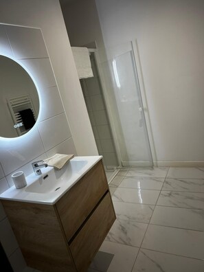 A modern bathroom with a white sink, wooden vanity, a round mirror, and a glass shower enclosure. - Cosy apartment ideally located in the center of Corsica, close to hiking and beaches. (PIEDIGRIGGIO)