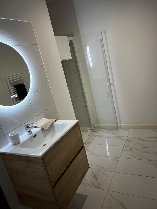 Ein modernes Badezimmer mit einem weißen Waschbecken, einer Holzausstattung, einem runden Spiegel und einer Dusche mit Glastrennwand.