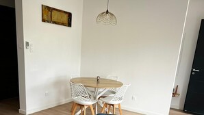 A living room with a blue sofa, a dining table with white chairs, a hanging light fixture, and a remote control on a yellow side table. - Cosy apartment ideally located in the center of Corsica, close to hiking and beaches. (PIEDIGRIGGIO)