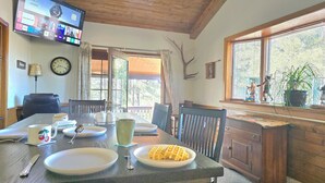A dining area with a wooden table, chairs, a TV mounted on the wall, a clock, and a window with a view of trees. - Munds Park Basecamp Cabin (Sedona)