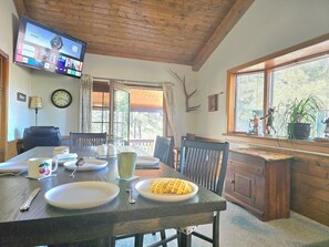 A dining area with a wooden table, chairs, a TV mounted on the wall, a clock, and a window with a view of trees. - Munds Park Basecamp Cabin (Sedona)