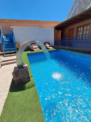 Uma piscina com um recurso aquático, uma escadaria e um edifício com varanda.