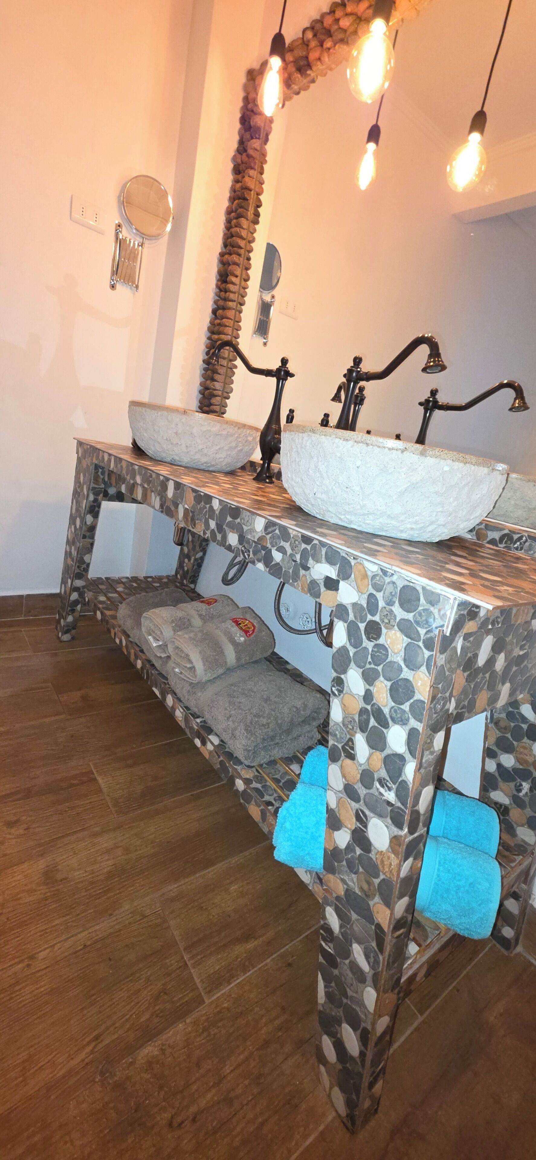 Un baño con tocador de mosaico de piedra, un espejo redondo y una lámpara con bombillas expuestas.