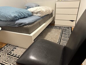 Uma cama com almofadas estampadas, uma mesa de cabeceira branca, uma cadeira preta e um tapete padronizado.