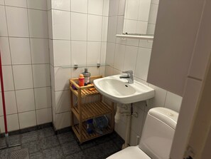 Um banheiro pequeno com azulejos brancos, uma prateleira de madeira com itens de higiene pessoal, uma pia branca e um vaso sanitário.