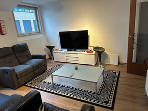 Un soggiorno con divano, tavolino, tappeto a motivi decorativi, televisore e una finestra vista su un edificio.