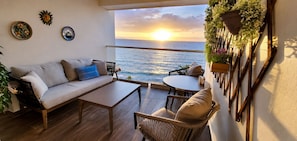 Ein Balkon mit einer Couch, einem Tisch und Stühlen mit Blick auf das Meer bei Sonnenuntergang.