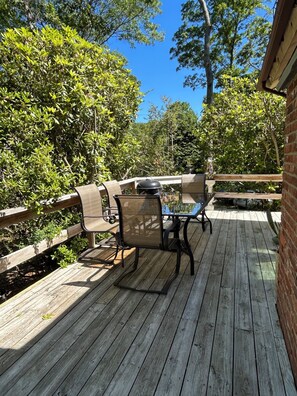 Una terraza de madera con mobiliario exterior, una parrilla y abundante vegetación.