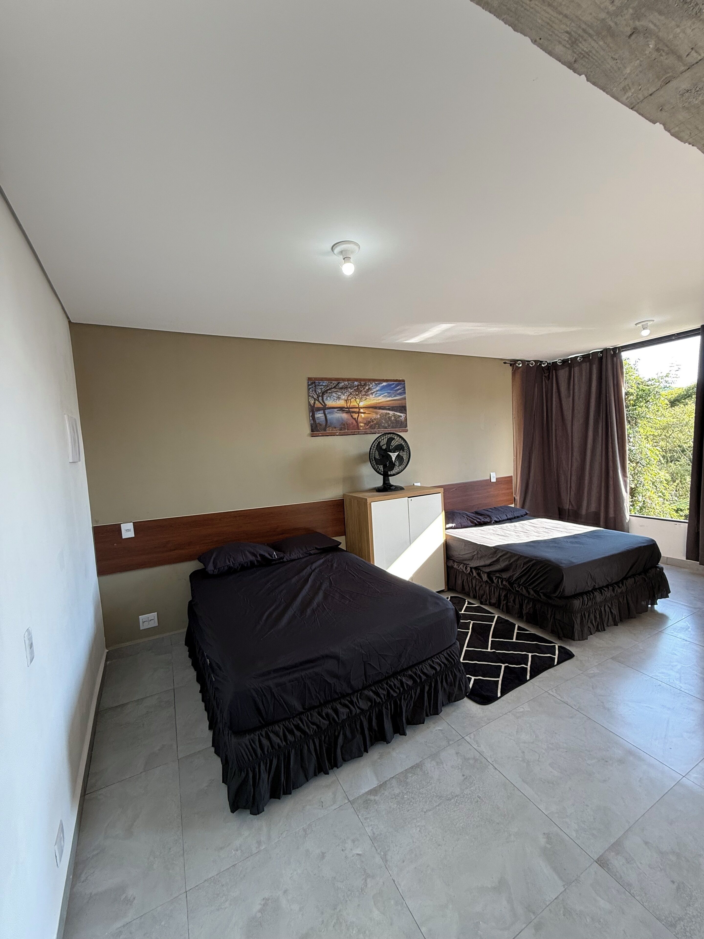 A bedroom with two beds, a desk, a fan, a painting, and a view of greenery.