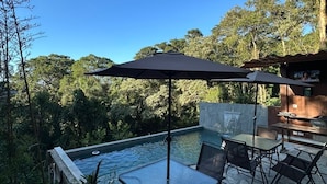 An outdoor pool area with a glass table and chairs, surrounded by stone flooring and a view of the forest. - House with infinity pool in Serra de Cantareira (Caieiras)