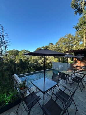 Ein Poolbereich im Freien mit einem Glastisch und Stühlen, umgeben von Steinböden und Blick auf den Wald.