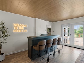 A modern kitchen with a bar area, wooden ceiling, and a 'WELCOME TO THE GOOD LIFE' sign.