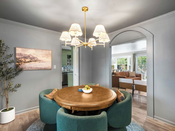 A dining area with a round wooden table, green upholstered chairs, a chandelier, and a view into the kitchen.