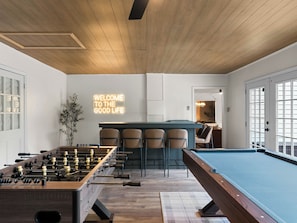 A room with a pool table, foosball table, bar area, and a 'WELCOME TO THE GOOD LIFE' sign.