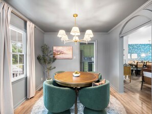 Una sala da pranzo con un tavolo rotondo, sedie rivestite in verde, un lampadario e vista sulla cucina attraverso una porta aperta.
