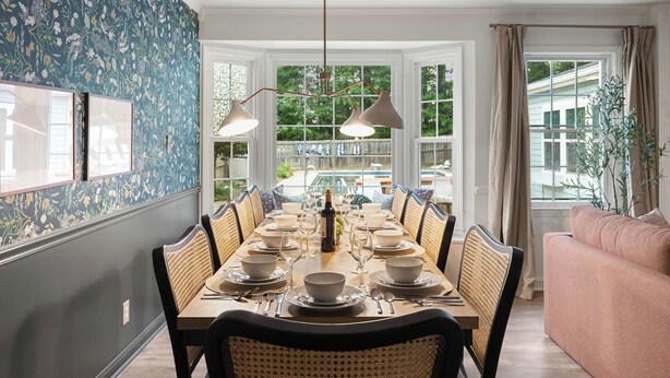 Un comedor con una larga mesa dispuesta para seis comensales, rodeada de sillas de mimbre, un candelabro y vistas al patio trasero a través de amplios ventanales.