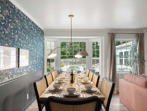 A dining room with a long table set for six, flanked by wicker chairs, a chandelier, and a view of the backyard through large windows. - Near Lake Keowee - Pool - Gameroom (Seneca)