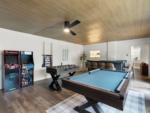 A pool table, foosball tables, and a welcome sign in a room with wooden flooring and a ceiling fan.