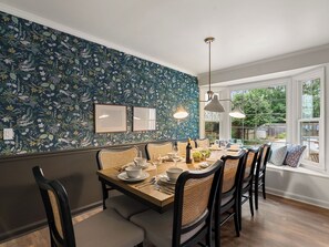 Un comedor con papel tapiz floral, una mesa de madera preparada para seis personas y un gran ventanal con vistas al exterior.