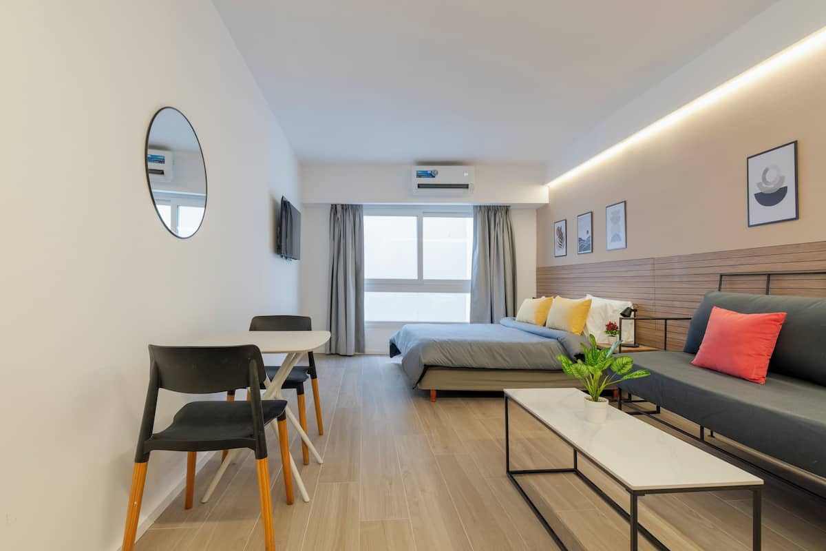 Ein modernes Hotelzimmer mit Bett, Sofa, Esstisch mit Stühlen, Spiegel und einer Zimmerpflanze.