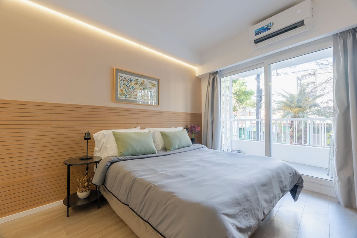 Ein modernes Schlafzimmer mit einem großen Bett, einem Nachttisch mit Lampe, einer Wandklimaanlage und einem großen Fenster mit Vorhängen.