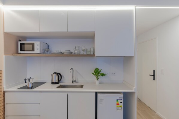 Une cuisine compacte avec des meubles blancs, un four à micro-ondes, une bouilloire, un évier et une petite plante en pot.