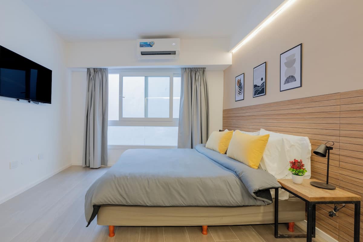 Ein modernes Schlafzimmer mit Bett, Nachttisch mit Lampe, einem an der Wand montierten Fernseher und einem Fenster mit Vorhängen.