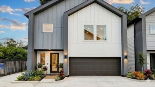 Et moderne hus i to etager med garage, en forhave med planter og en klar himmel.