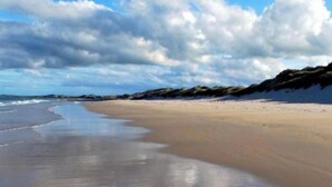 Uma praia com extensa faixa de areia, ondas suaves e céu parcialmente nublado.