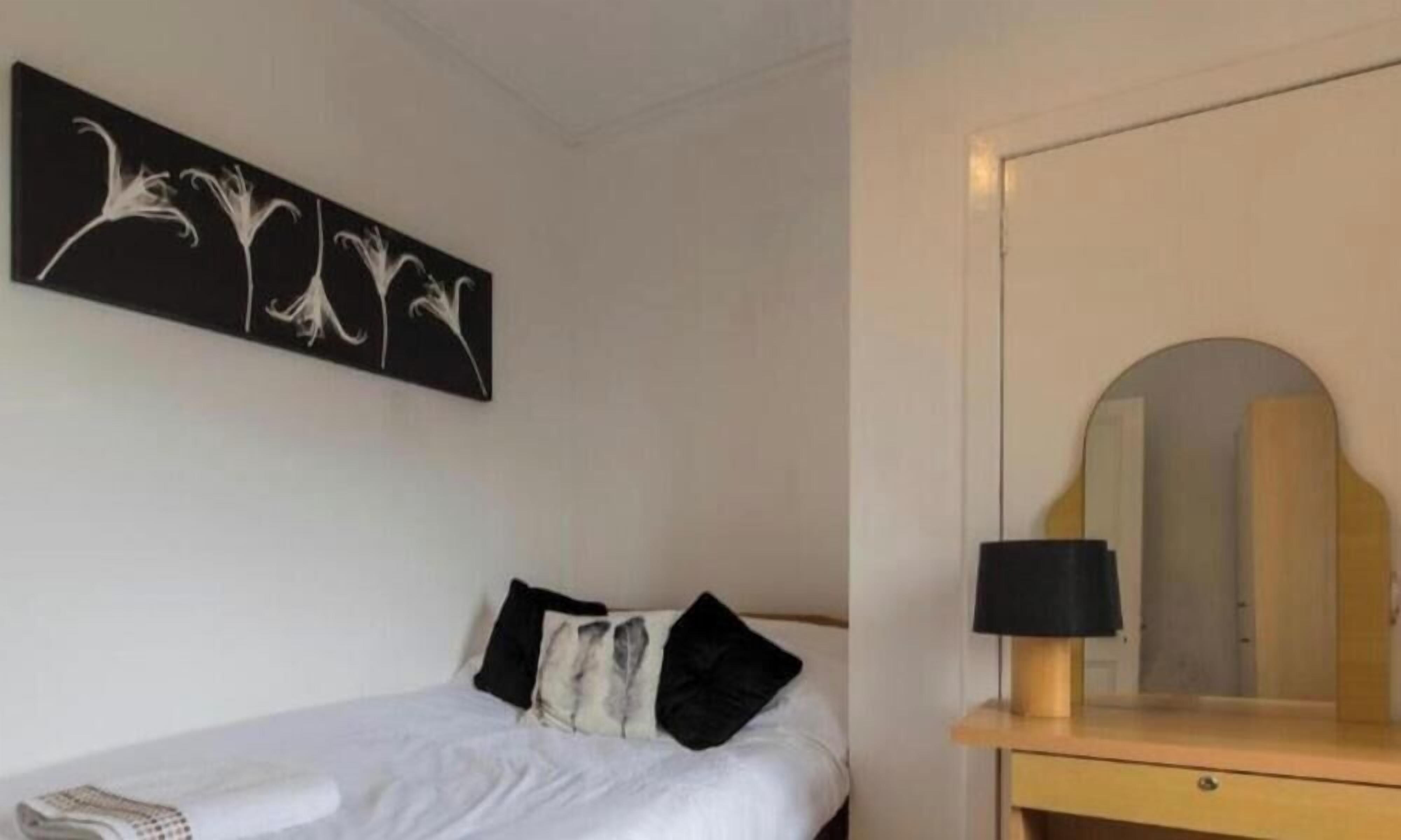 Ein Hotelzimmer mit einem Bett, einem Nachttisch mit Lampe, einem Spiegel und einem an der Wand hängenden Kunstwerk.