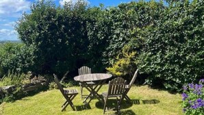 Um jardim com mesa e cadeiras de madeira, cercado por vegetação e flores.