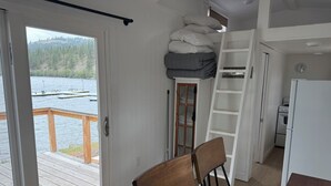 Una habitación con puerta de vidrio corrediza que da a una terraza de madera, un altillo con espacio de almacenamiento, una zona de cocina y vista a un lago.