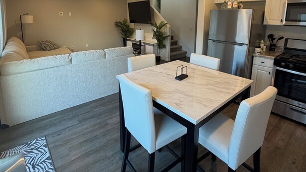 A modern kitchen with white cabinets, a stainless steel refrigerator, and a dining area with a marble table and white chairs. - Verano Gateway (Cathedral City)
