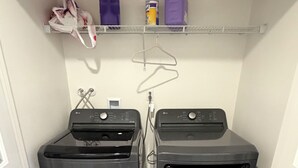 A laundry room with two front-loading washers and dryers, a shelf with cleaning supplies, and a hanging rack. - Verano Gateway (Cathedral City)