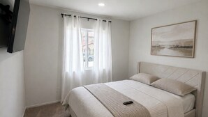A bedroom with a bed, a flat-screen TV mounted on the wall, a window with curtains, and a painting on the wall. - Verano Gateway (Cathedral City)
