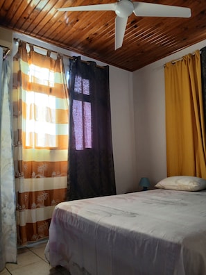 Ein Schlafzimmer mit einem Bett, Deckenventilator, einem Fenster mit Vorhängen und einem Nachttisch mit Lampe.
