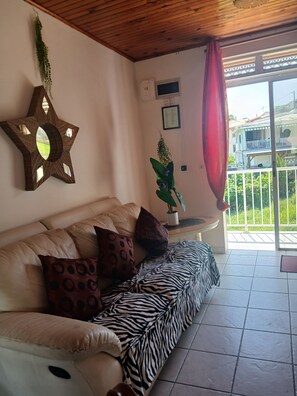 Ein Wohnzimmer mit einem sternförmigen Spiegel, einer Couch mit gemusterten Kissen, einer Zimmerpflanze und Blick auf einen Balkon.