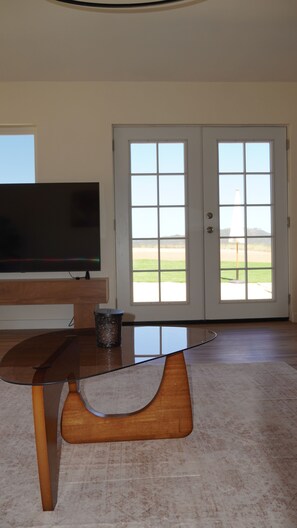 A room with a television on a wooden stand, a glass coffee table, and a door leading to a view of a field. - 4-bedroom ranch house in peaceful Malibu with ocean and mountains view (Malibu)
