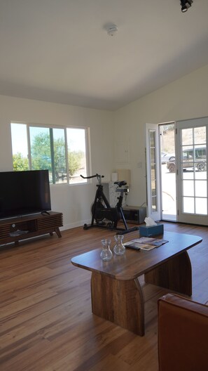 A spacious living room with a wooden floor, a large flat-screen TV, a standing exercise bike, and a dining table with two clear vases. - 4-bedroom ranch house in peaceful Malibu with ocean and mountains view (Malibu)
