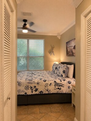 Ein Schlafzimmer mit einem Bett, einem Deckenventilator, einem Fenster mit Jalousien, einem Gemälde und einer Tür.
