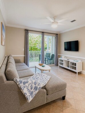 Ein Wohnzimmer mit Sofa, Couchtisch, Fernseher und einem Balkon mit Blick auf Bäume.