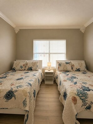 Une chambre avec deux lits, une fenêtre avec des stores, une table de chevet avec une lampe et un plancher en bois.