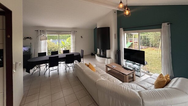 Ein modernes Wohnzimmer mit Sofa, Esstisch, Fernseher und großen Fenstern.