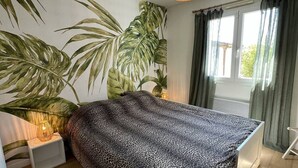 Ein Schlafzimmer mit Bett, einer Hängeleuchte, einem Fenster mit Vorhängen und einer großen Pflanze.