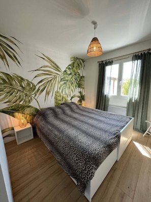 Ein Schlafzimmer mit Bett, einer Hängeleuchte, einem Fenster mit Vorhängen und einer großen Pflanze.