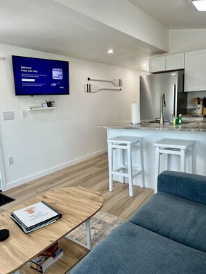 A modern kitchen with a flat-screen TV, white bar stools, a grey sofa, and a wooden coffee table. - GOTL- Sun Retreats/Heated Pool/Free Golf Cart (Geneva)