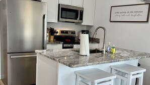 Una cocina moderna con un refrigerador de acero inoxidable, un microondas y un horno empotrado. Hay dos taburetes blancos y una encimera de mármol.